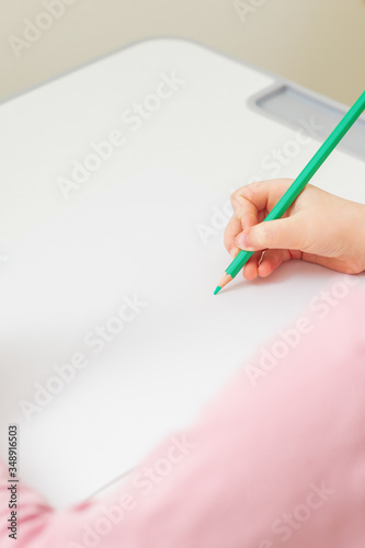 Vertical image of girl's right hand holding pencil over a white blank sheet of paper on a white desk. Copy space for text. Mockup. Selective focus.