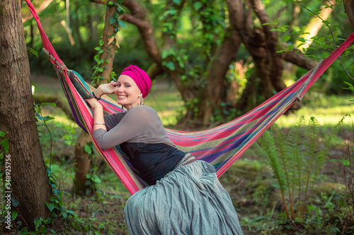 A woman in a purple turban is smiling. sitting in a hammock in the forest. touches the face with a hand.