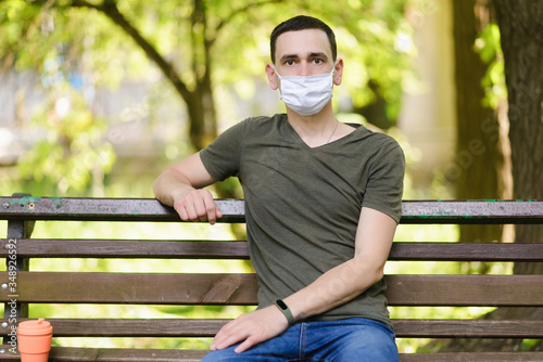A young man in a medical mask sits in a park. Quarantine restrictions are caused by coronavirus