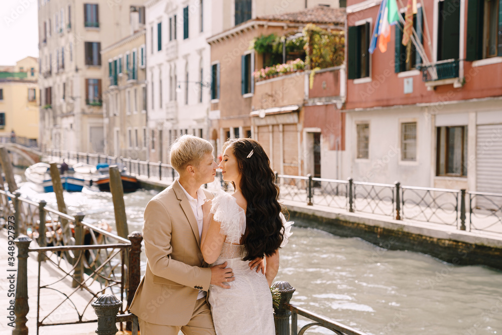 Italy wedding in Venice. Newlyweds stand embracing on the banks of the Venice Canal. The groom hugs the bride by the waist. White wedding dress with small beautiful train and sand-colored men's suit.