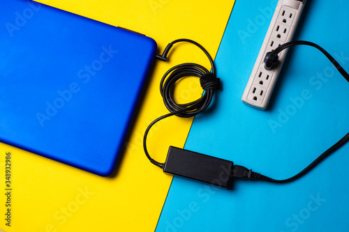 Laptop, charger and Surge Protector Power over blue yellow background. Battery life concept. topic of electric energy in travel. mobility concept. digital nomad concept. job online concept.