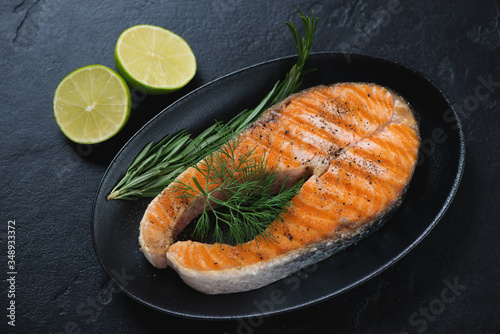 Grilled salmon steak with fresh dill, rosemary and lime served on a black plate, horizontal shot on a black stone surface