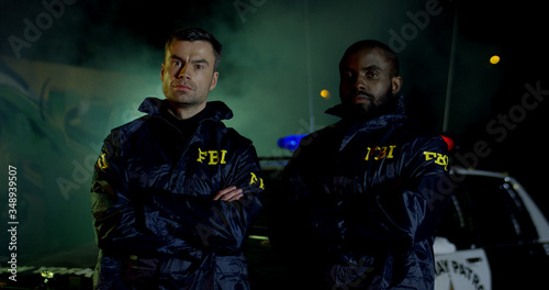 Portrait of Two police officers with serious faces looking at camera. FBI agents work at the scene at night, police car with lights on background photo