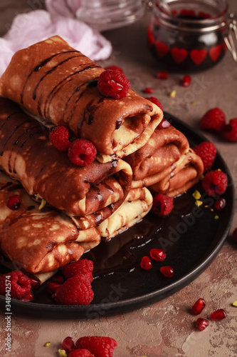 Sweet pancakes with cottage cheese filling, poured with chocolate topping and raspberries. Decorated with fresh pomegranate and raspberry, with a jar of chocolate on a dark background to pancake week