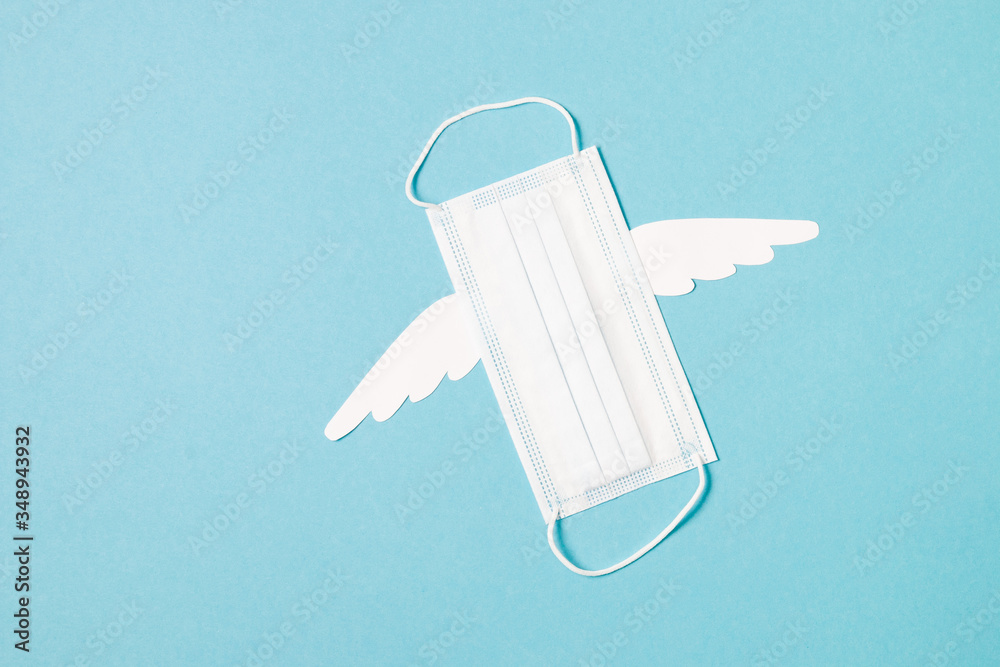 medical protective mask with white paper wings on a light blue background, copy space, coronavirus pandemic concept, cancellation of flights and travel, quarantine 2020. medical protective mask with white paper wings on a light blue background, copy space, coronavirus pandemic concept, cancellation of flights and travel, quarantine 2020.