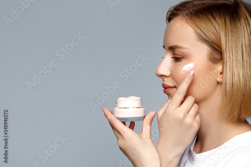 Photos Close up woman holding bottle of cream in hand and apply it on face with finger