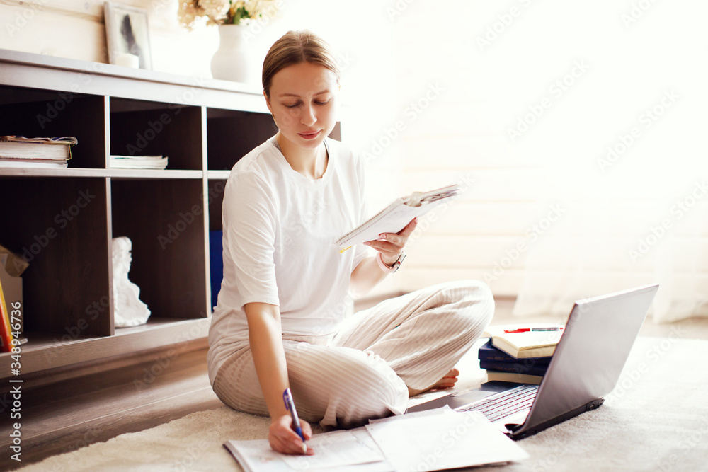 Woman sitting on floor on carpet at home in cozy homewear, remote working or online studying, with papers, notebook and computer, writing and reading, at sunny day. Online bussiness or education Woman sitting on floor on carpet at home in cozy homewear, remote working or online studying, with papers, notebook and computer, writing and reading, at sunny day. Online bussiness or education