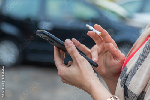 Female hands holding a mobile phone and having a lit cigarette between them, which is smoking and a car in the background