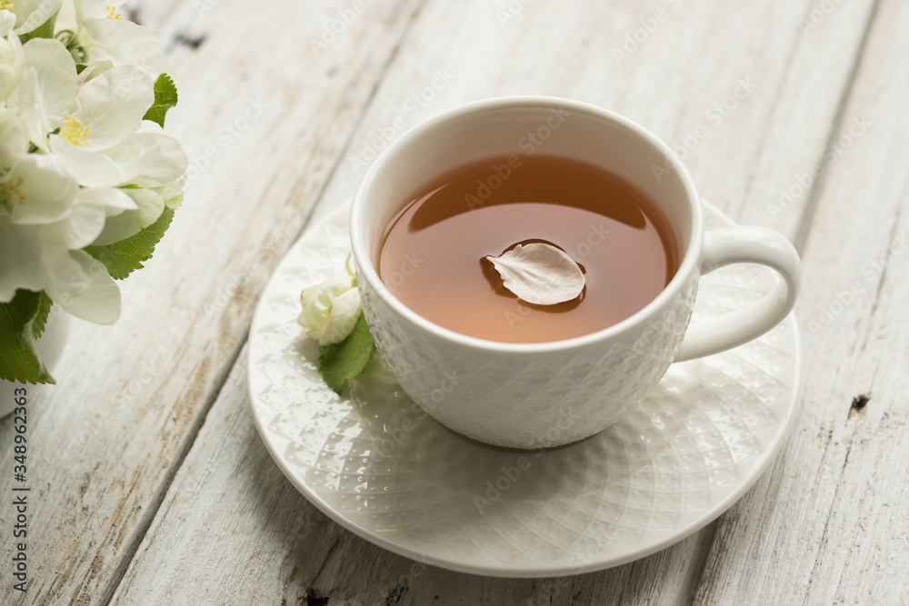 A cup of tea and a branch of a blossoming apple tree on the table. Flower petals in a drink A cup of tea and a branch of a blossoming apple tree on the table. Flower petals in a drink