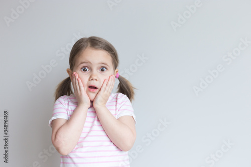 Portrait of a white girl 4 years old. She is very surprised, her mouth is open, her hands on her cheeks. Attractive. Discounts, sale. Grey background. Incredible. Place for text, layout.
