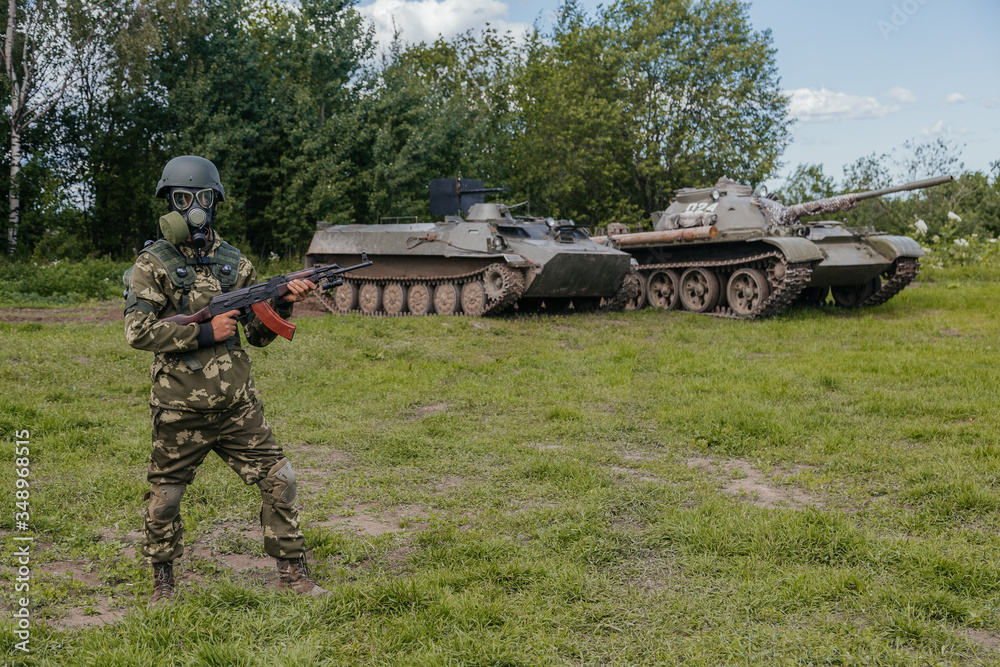 A soldier in a gas mask with a Kalashnikov rifle against the background of armored vehicles.