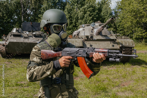 A soldier in a gas mask with a Kalashnikov rifle against the background of armored vehicles.