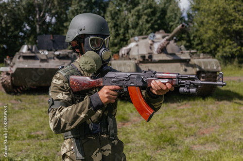A soldier in a gas mask with a Kalashnikov rifle against the background of armored vehicles.