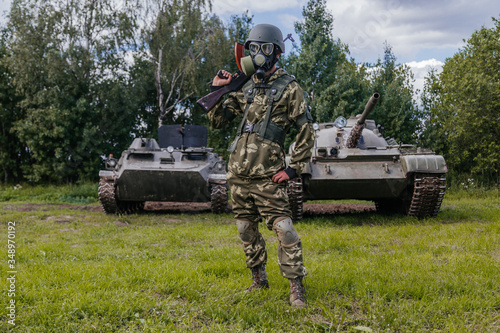 A soldier in a gas mask with a Kalashnikov rifle against the background of armored vehicles.