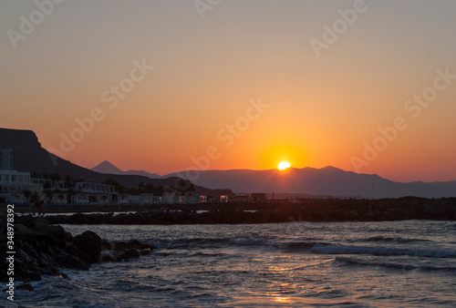 Sunset over the sea in Kokkini Hani, Crete, Greece. The sun disappears behind the mountain. Scenic seaside landscape in the evening.