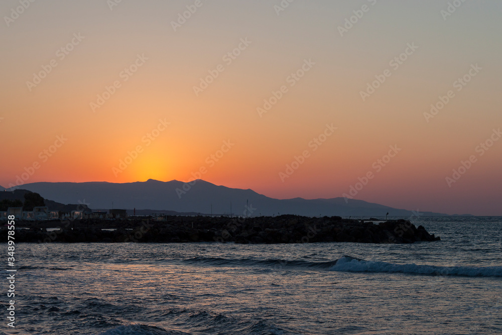 Sunset over the sea in Kokkini Hani, Crete, Greece.  The sun disappears behind the mountain. Scenic seaside landscape in the evening.