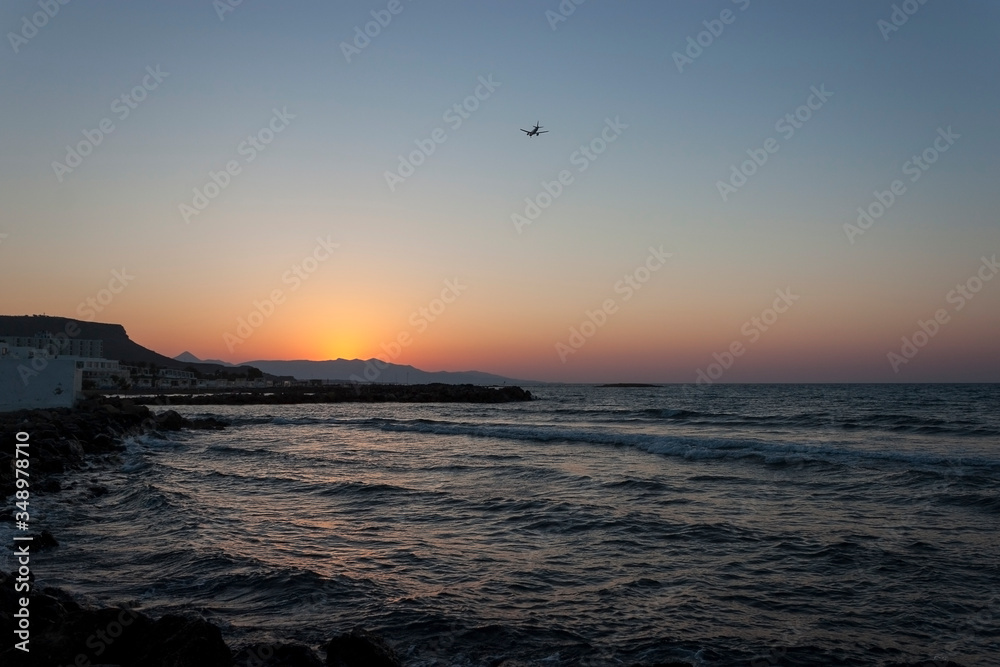 Sunset over the sea in Kokkini Hani, Crete, Greece. The sun disappears behind the mountain. A plane is flying in the sky. Scenic seaside landscape in the evening. Sunset over the sea in Kokkini Hani, Crete, Greece. The sun disappears behind the mountain. A plane is flying in the sky. Scenic seaside landscape in the evening.