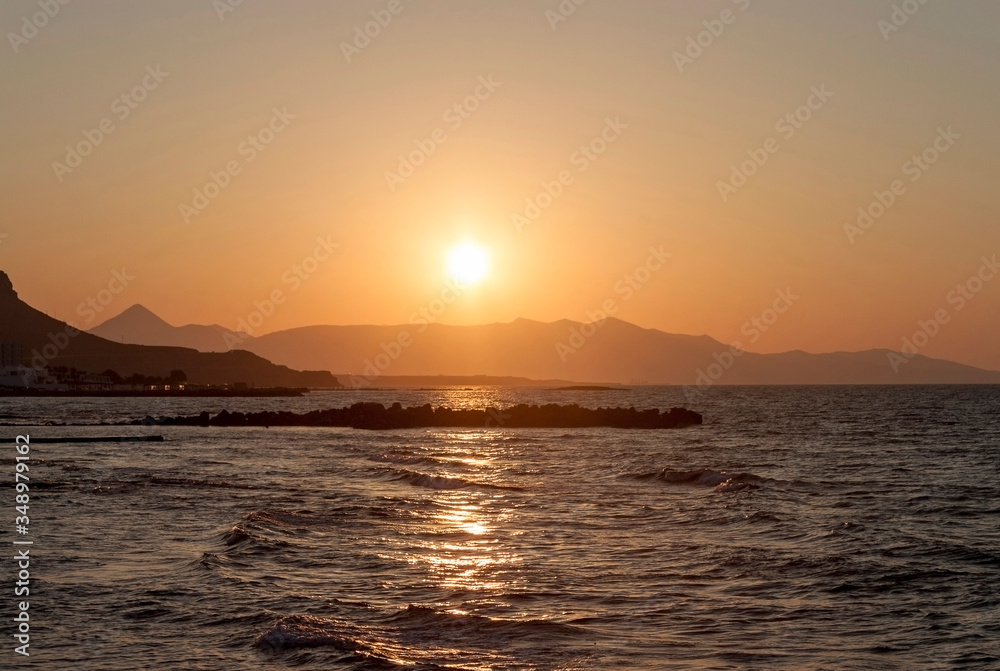 Sunset over the sea in Kokkini Hani, Crete, Greece.  The sun disappears behind the mountain. Scenic seaside landscape in the evening.
