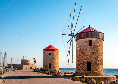 Situated on the long wave breaker at Mandraki harbour, Rhodes, Greece, stand these medieval windmills. The fortress and the lighthouse of Agios Nikolaos (Saint Nicholas) is in the background.