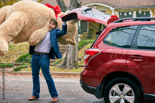 Man grabs a valentines day gift out of the trunk of his car, surprising his partner with a large teddy bear.