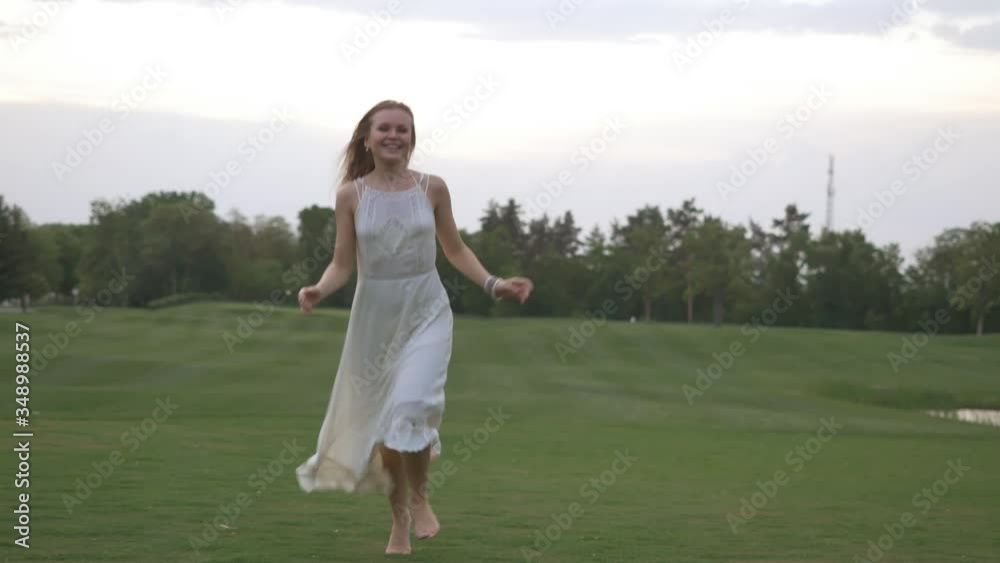Beautiful blonde girl in a white dress runs and dances barefoot on the green grass in summer day Beautiful blonde girl in a white dress runs and dances barefoot on the green grass in summer day