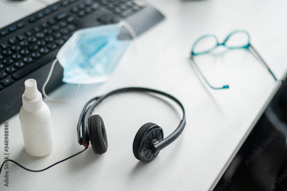 Headset medical mask and antiseptic on the desktop of a call center operator desk. Glasses near the keyboard at the workplace in the office. The concept of safe work during the coronavirus epidemic. Headset medical mask and antiseptic on the desktop of a call center operator desk. Glasses near the keyboard at the workplace in the office. The concept of safe work during the coronavirus epidemic.
