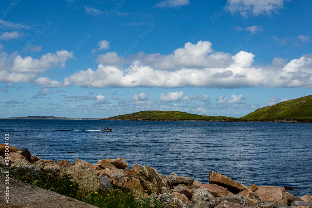 Motorboat sailing in the calm water with rolling hills with green grass in the background viewed from the shoreline on Inishbofin Island, sunny and calm spring day in County Galway, Ireland Motorboat sailing in the calm water with rolling hills with green grass in the background viewed from the shoreline on Inishbofin Island, sunny and calm spring day in County Galway, Ireland