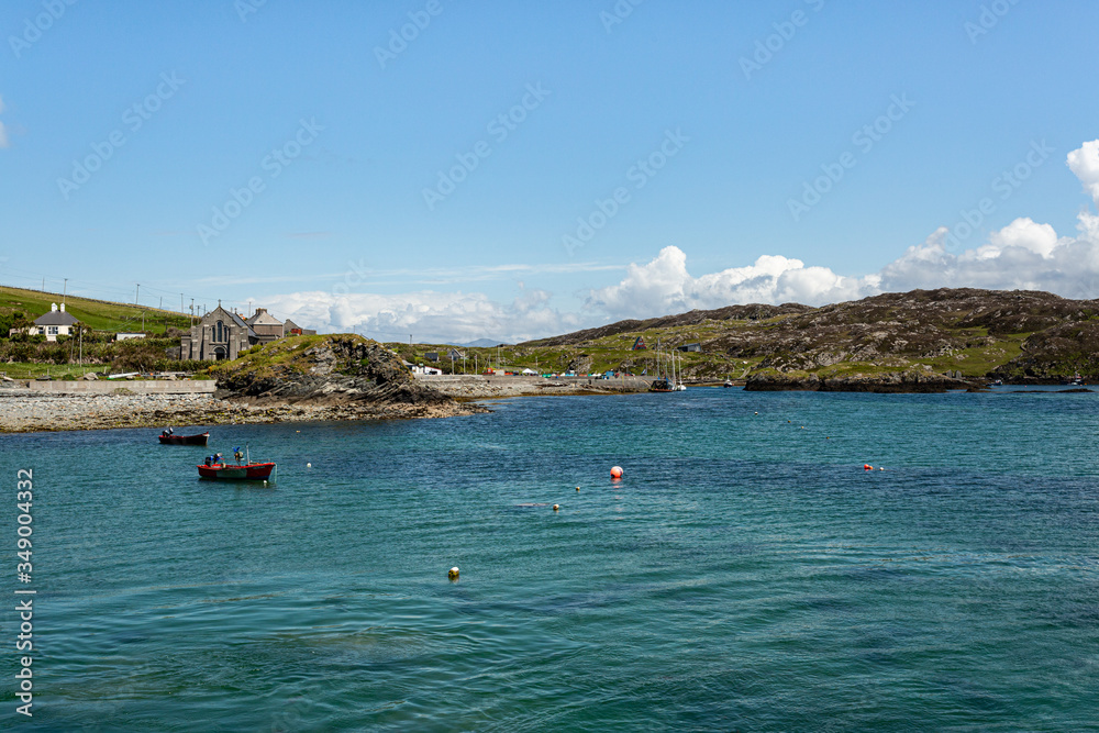 Boats and buoys in the bay, the rocky shoreline with its small harbor, its church on Inishbofin Island, sunny spring day with blue sky in County Galway, Ireland