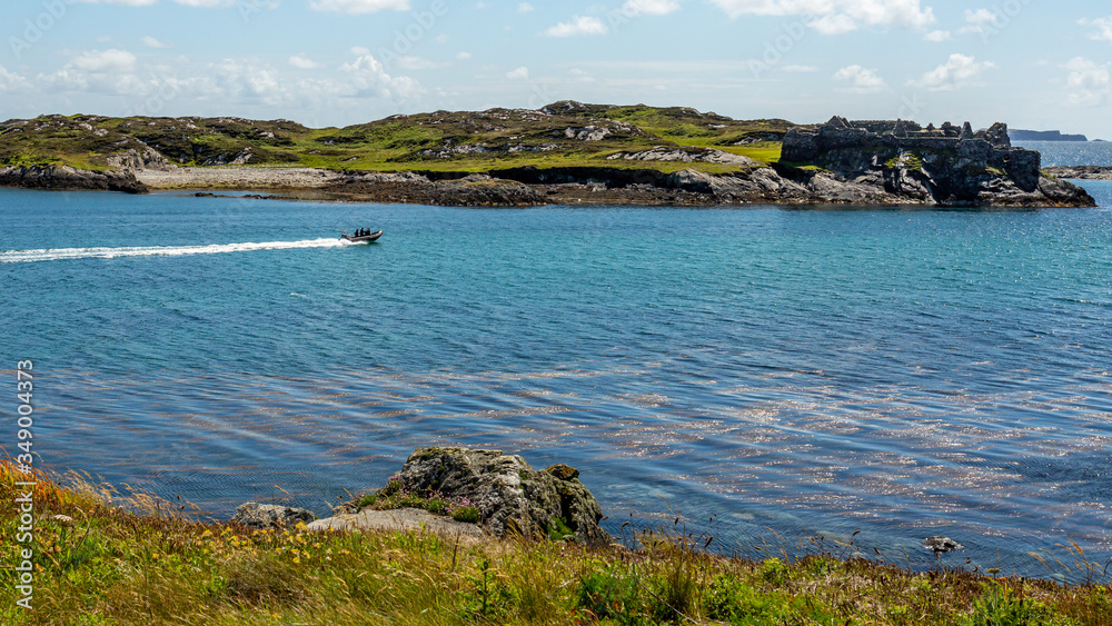Bay with a motorboat sailing with the ruins of Cromwell’s Barracks in the background on Inishbofin Island, sunny and calm spring day in County Galway, Ireland