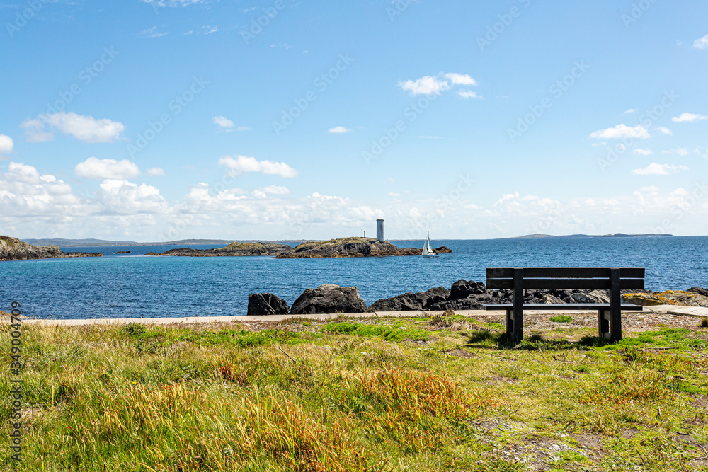 Wooden bench on the coastal shoreline in Inishbofin or White Cow Island with the lighthouse on Gun Rock and a sailboat in the background, sunny spring day in County Galway, Ireland
