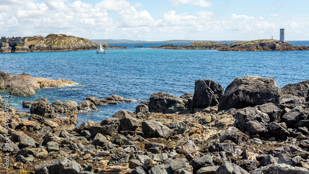 Rocky shoreline with Cromwell's Barracks, a sailboat and the lighthouse on Gun Rock in the background on Inishbofin or White Cow Island, sunny spring day in County Galway, Ireland