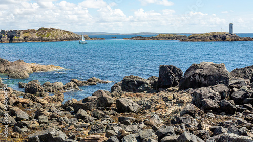 Rocky shoreline with Cromwell's Barracks, a sailboat and the lighthouse on Gun Rock in the background on Inishbofin or White Cow Island, sunny spring day in County Galway, Ireland