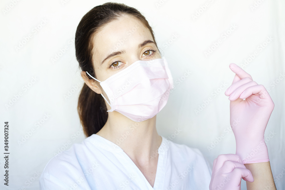 A woman in a pink protective mask adjusts her pink gloves. The girl stands on a light background in white clothes. The concept of suppression of coronavirus
