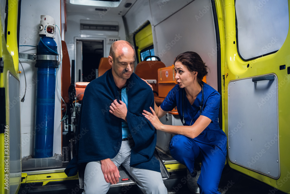 Female paramedic talking to an injured man trying to comfort him, the man is sitting on the edge of an ambulance car in a blanket Female paramedic talking to an injured man trying to comfort him, the man is sitting on the edge of an ambulance car in a blanket