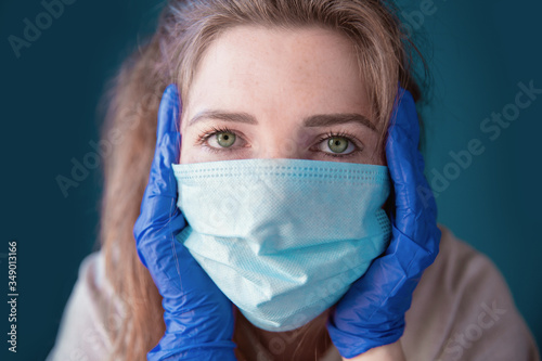 Young woman wearing protective gloves and face mask inside a home in quarantine looking bored and sad, for Covid-19 Coronavirus, with blue background