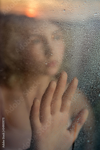 Silhouette of a girl behind a glass with raindrops. Reflection of sunset in the glass. Conceptual photography: the theme of loneliness,self-isolation, fencing off.