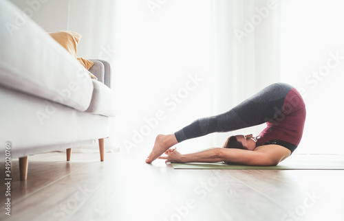 Young graceful Woman enjoying morning yoga exercises doing Halasana pose at home living room near the big window. Active people, body care and healthy lifestyle concept. photo