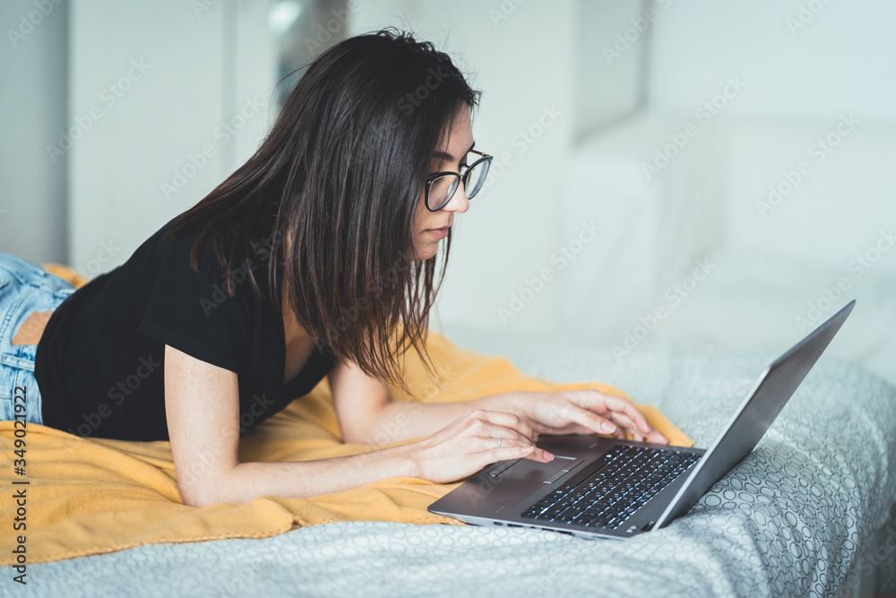 Pretty brunette woman wearing eyeglasses and working on laptop computer. Concentrated female lying in bed at home writing and using .notebook keyboard touchpad. Freelance lifestyle and work from home