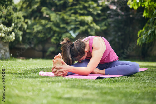 Attractive smiling caucasian brunette doing yoga in her backyard. Seated Three Limbed Forward Bend yoga pose.