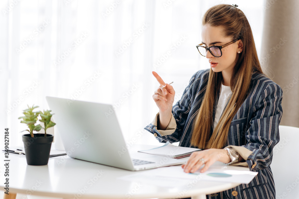 Online business meeting. Attractive business woman with glasses and stylish formal clothes communicates online through video call on a laptop with her colleagues on her workplace at home Online business meeting. Attractive business woman with glasses and stylish formal clothes communicates online through video call on a laptop with her colleagues on her workplace at home