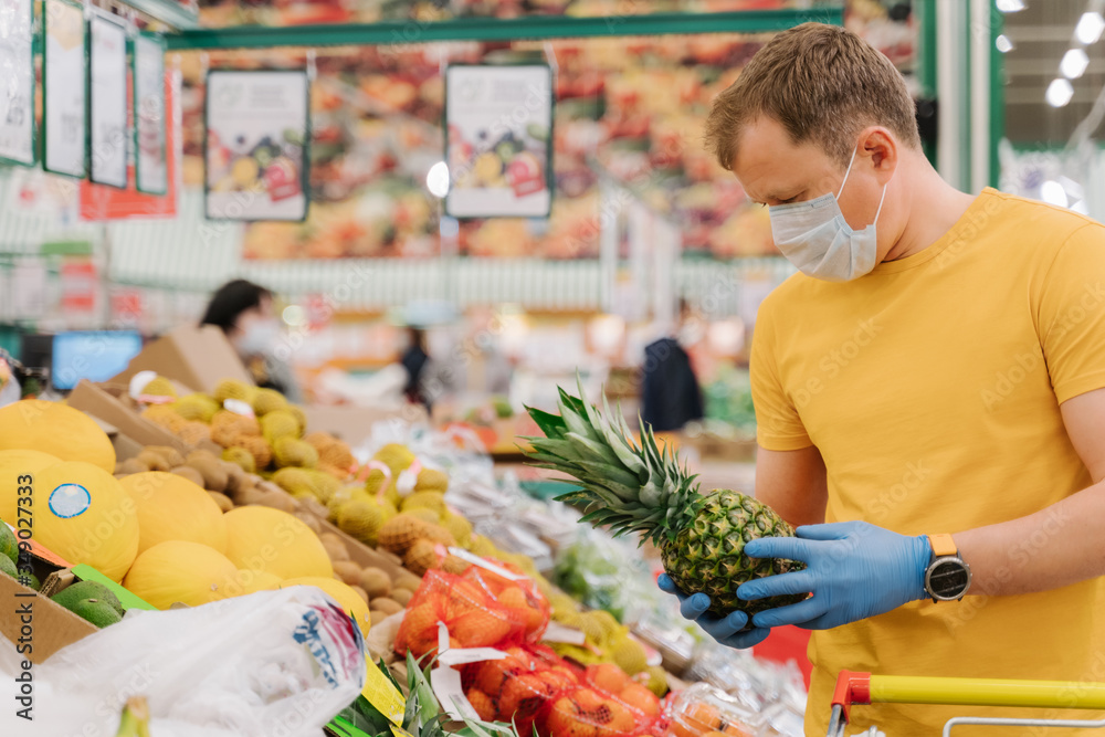 Obraz premium Horizontal shot of man selects pineapple in store wears medical mask and gloves buys fresh fruit during coronavirus outbreak or virus pandemic poses in grocery department. Protection, prevent measures