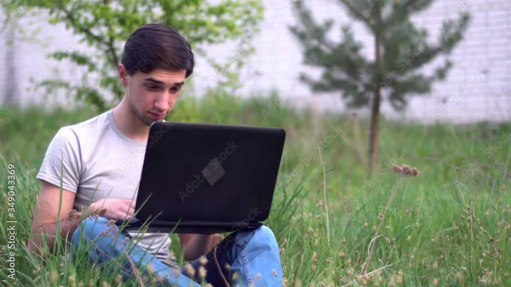 A young caucasian man sits on the grass near the house and typing on a laptop. Remote work concept A young caucasian man sits on the grass near the house and typing on a laptop. Remote work concept