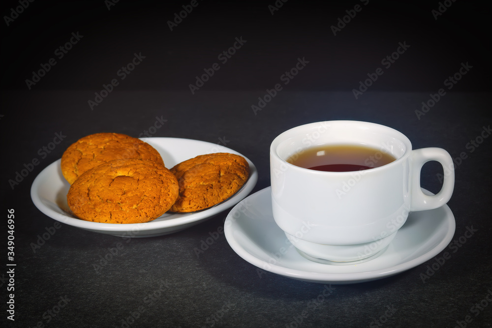 White cup with black tea and oatmeal cookies on a white saucer.