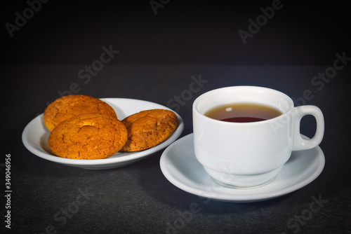 White cup with black tea and oatmeal cookies on a white saucer.
