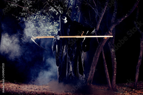 Portrait of grim reaper, the angel of death ( Azrael ) lurking through the woods at night surrounded with mist, smoke in a mystical forest holding his shythe. photo