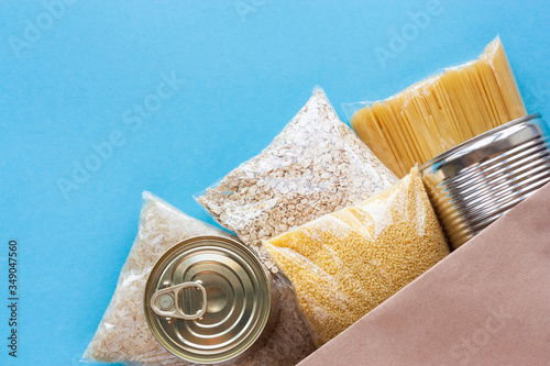 Various grocery products in packages top view. Pasta, cereals and canned food in paper bag on blue background. Food delivery, donation or stock provision concept. Copy space.