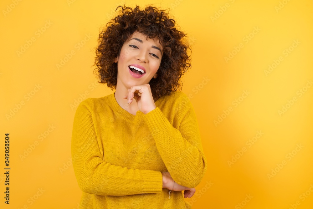Optimistic Arab woman, curly hair, keeps hands partly crossed and hand under chin, looks at camera with pleasure, wears yellow sweater, isolated over yellow background. Happy emotions concept. Optimistic Arab woman, curly hair, keeps hands partly crossed and hand under chin, looks at camera with pleasure, wears yellow sweater, isolated over yellow background. Happy emotions concept.