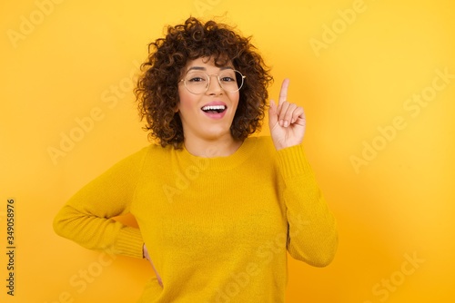 Young pretty arabian girl wearing yellow sweaterholding finger up having idea and posing on yellow background.