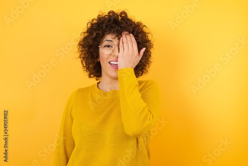 Young beautiful brunette woman wearing yellow sweater over yellow isolated background covering one eye with her hand, confident smile on face and surprise emotion. © Roquillo