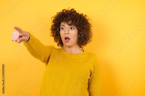 Young beautiful woman wearing casual yellow sweater over isolated yellow background Pointing with finger surprised ahead, open mouth amazed expression, something on the front.